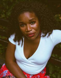 I.S Jones looks up at the camera while sitting outdoors among greenery. She wears a white wrap-style top and a red floral skirt.