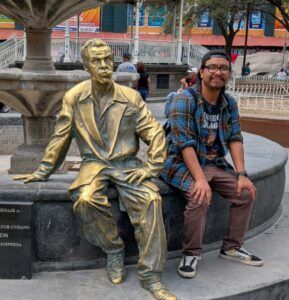 Photo of Miguel Mendez sitting at a fountain next to a gold statue